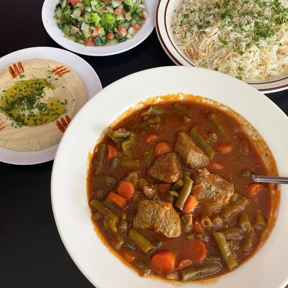 Mediterranean meal featuring beef and vegetable stew, herb-topped rice, chopped salad, and hummus arranged on white plates.